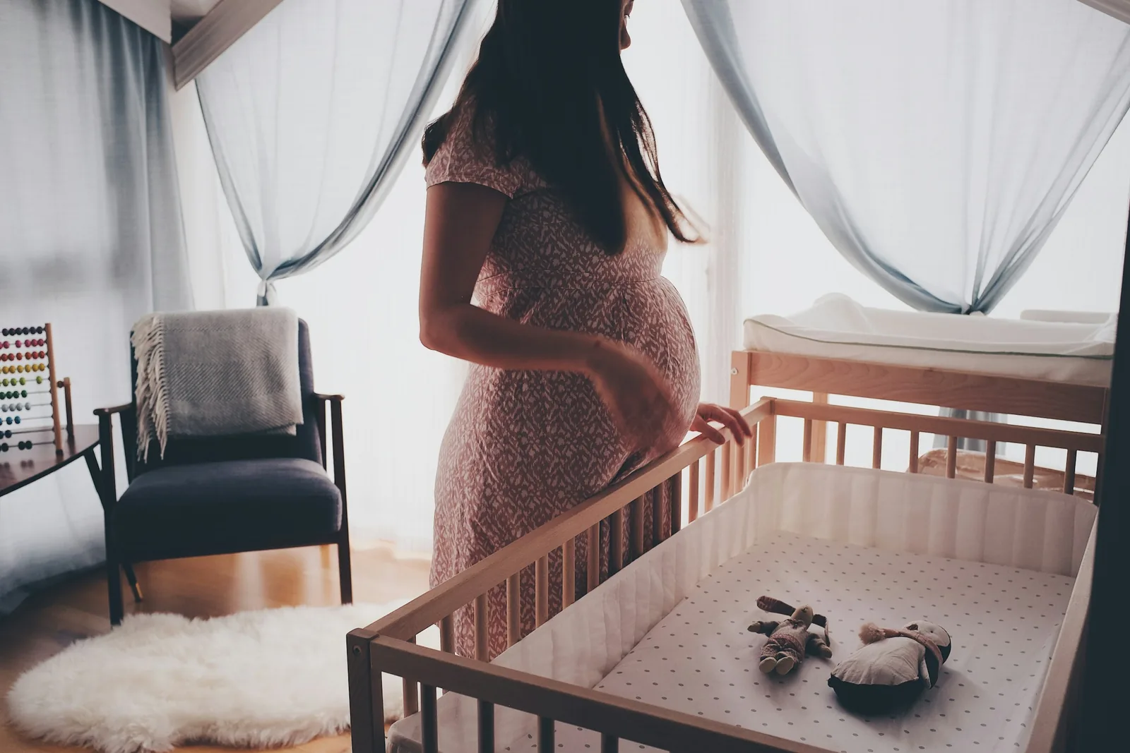 Pregnant woman standing in a sunlit nursery beside a crib